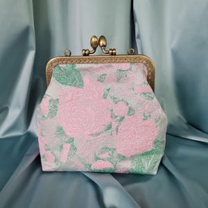 May include: A small, floral patterned clutch purse with a brass clasp. The purse is made of a soft, textured fabric with a pink and green floral design.