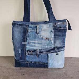 Puede incluir: Bolso tote de mezclilla patchwork con diferentes tonos de azul. El bolso tiene cierre de cremallera y asas de mezclilla. Cuenta con parches de bolsillos, trabillas y diversas texturas de mezclilla. Diseñado para transportar objetos.