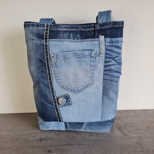 Könnte beinhalten: Eine Denim-Tragetasche aus verschiedenen Jeansblau-Tönen. Die Tasche hat eine hellblaue Jeanstasche auf der Vorderseite, einen silbernen Knopf und eine kontrastierende dunkelblaue Jeansbahn. Die Griffe sind aus demselben Denim-Material.