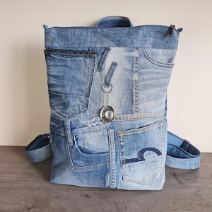 Op de afbeelding: Een rugzak gemaakt van hergebruikte blauwe spijkerstof met meerdere zakken, zichtbare naden en een ritssluiting aan de bovenkant. Een kleine zilverkleurige cowboyhoed-bedel hangt aan een metalen ring aan de voorkant van de tas.