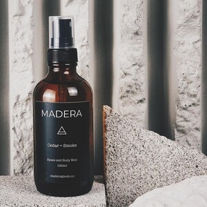 Pode incluir: Uma garrafa de vidro marrom com um rótulo preto que diz "MADERA" e "Cedar + Smoke". A garrafa contém um spray para o espaço e o corpo e tem uma capacidade de 120 ml. A garrafa está sobre uma superfície cinza texturizada.