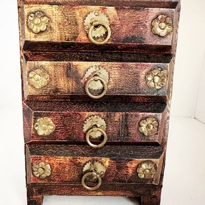 May include: A small wooden chest with five drawers. Each drawer has a brass ring pull and a decorative brass flower design. The chest is painted in shades of brown and gold.