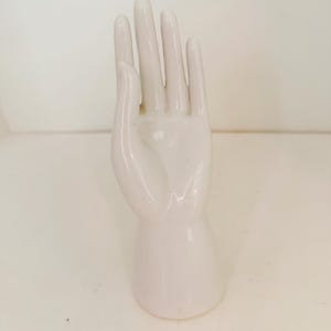 May include: A glossy white ceramic hand sculpture designed as a display stand. The upright hand features extended fingers and a smooth, reflective surface, standing on a cylindrical base that provides stability for the upright structure.