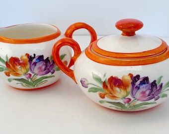 May include: A white ceramic creamer and sugar set with orange handles and floral designs. The flowers are painted in shades of orange, yellow, purple, and green.