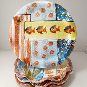 May include: A set of four ceramic plates with a scalloped edge. The plates feature a colorful, hand-painted design with orange fish, blue and orange flowers, and stripes.