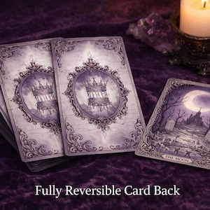 May include: A deck of rectangular tarot cards with a purple and white colour palette. The card backs feature an ornate frame surrounding a crown illustration. One card is face up, displaying a moonlit landscape with the text The Moon at the bottom.