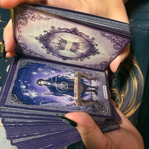 May include: A deck of tarot cards with purple edges and ornate borders. The top card features a crown inside a decorative frame. The exposed card displays an illustration of a figure titled The Magician, standing at a table with glowing energy.