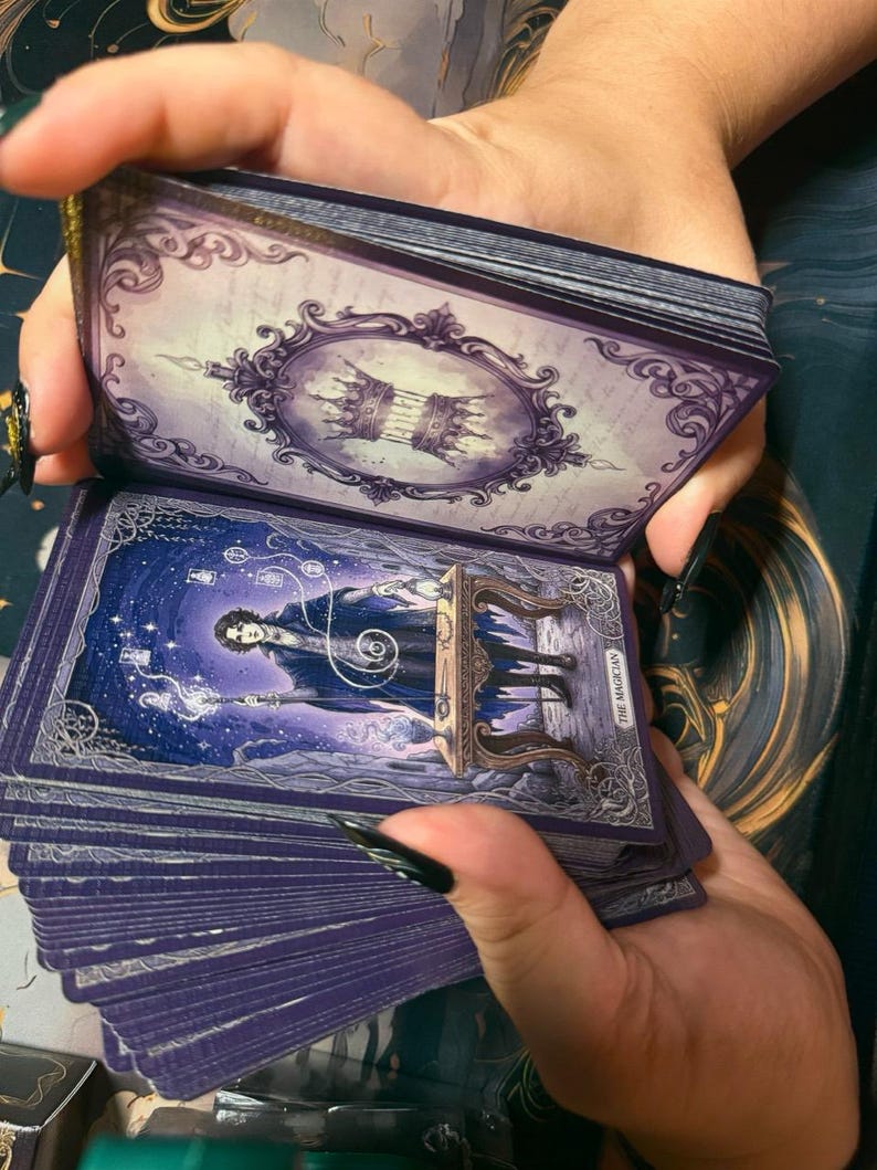May include: A deck of tarot cards with purple edges and ornate borders. The top card features a crown inside a decorative frame. The exposed card displays an illustration of a figure titled The Magician, standing at a table with glowing energy.