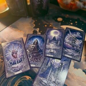 May include: A deck of tarot cards titled The Queens Madness with dark purple and blue illustrations. Visible cards include Three of Swords, Strength, The Moon, Knight of Swords, and Two of Cups, all featuring intricate fantasy artwork and text labels.