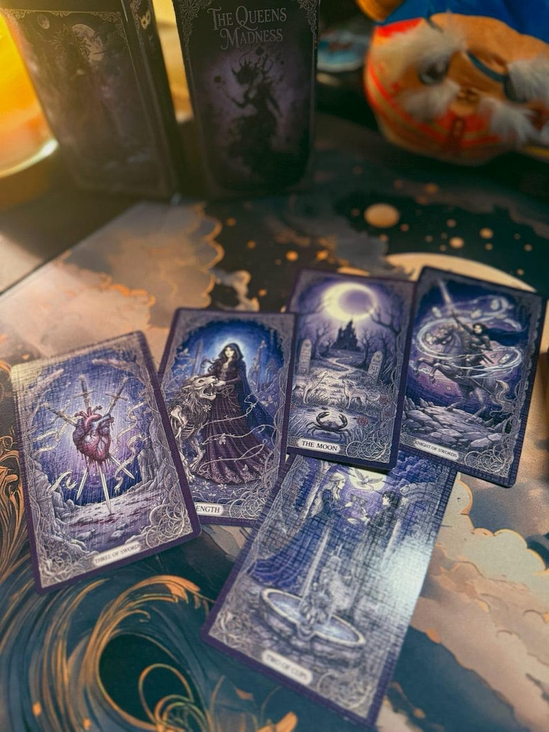 May include: A deck of tarot cards titled The Queens Madness with dark purple and blue illustrations. Visible cards include Three of Swords, Strength, The Moon, Knight of Swords, and Two of Cups, all featuring intricate fantasy artwork and text labels.