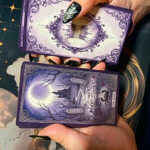 May include: A deck of tarot cards featuring a purple colour palette. The top card back displays an ornate frame with a crown motif. The bottom card face shows a moonlit landscape with a castle, a wolf, a dog, and a crab, labelled The Moon.