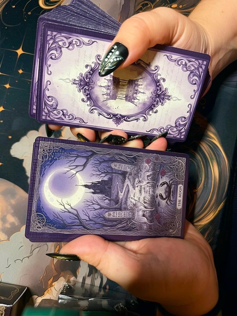 May include: A deck of tarot cards featuring a purple colour palette. The top card back displays an ornate frame with a crown motif. The bottom card face shows a moonlit landscape with a castle, a wolf, a dog, and a crab, labelled The Moon.