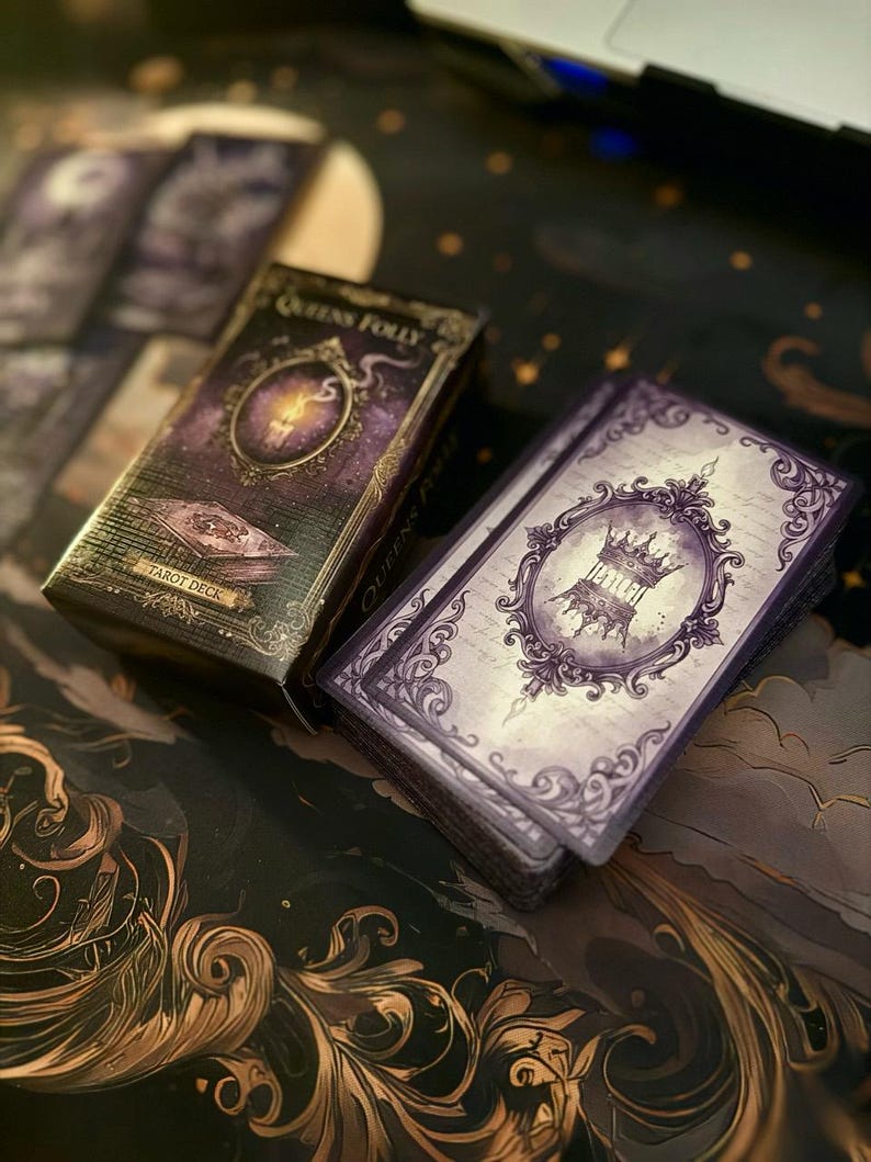 May include: A Queens Folly tarot deck featuring a rectangular box with a purple candle illustration and a stack of cards displaying a purple ornate frame with a crown graphic. The design includes dark purple and black tones with intricate scrollwork.