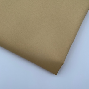 Op de afbeelding: Een gevouwen stuk tan stof, mogelijk katoen of een vergelijkbaar materiaal, met een subtiele geweven textuur. De stof heeft een effen, neutrale kleur en de randen zijn netjes afgesneden. De stof wordt gepresenteerd tegen een witte achtergrond.