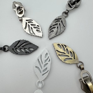Op de afbeelding: Close-up van vijf metalen ritstrekkers met blad-vormige bedels. De trekkers zijn in zilver, gunmetal, zwart, wit en messing tinten. De bladbedels hebben ingewikkelde uitgesneden ontwerpen, en de ritsen zijn aan de trekkers bevestigd.