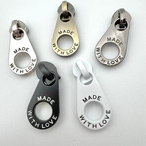 May include: Five metal zipper pulls in silver, gold, black, and white. Each pull has the words "MADE WITH LOVE" engraved around a central hole. The pulls are teardrop-shaped with a loop at the top.