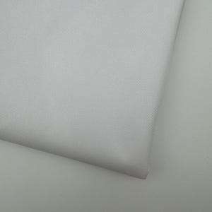 Op de afbeelding: Close-up van een opgevouwen stuk lichtgrijze stof. De stof heeft een subtiele textuur en lijkt een glad, geweven materiaal te zijn. De hoek van de stof is zichtbaar en toont de dikte en de manier waarop deze vouwt.