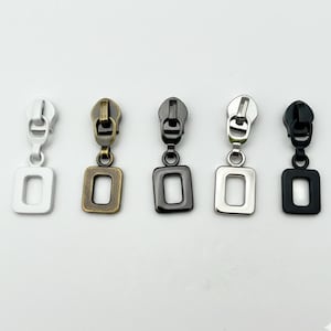 May include: Five metal zipper pulls in silver, antique brass, gunmetal, chrome, and black. Each pull has a rectangular loop attached to the zipper head. These pulls are used for clothing, bags, and other sewing projects.