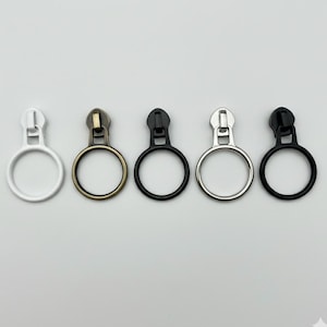 May include: Five metal zipper pulls in various colors: white, antique brass, black, silver, and black. Each pull features a circular ring for easy handling and a classic zipper design. Ideal for clothing and craft projects.