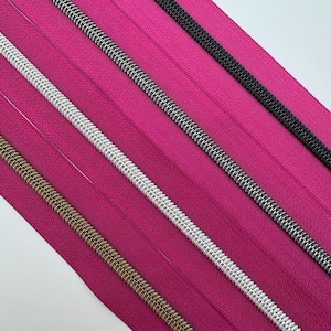 May include: Close-up of several zippers in various colors, including gold, silver, black, and a silver and black combination. The zippers are arranged diagonally across a bright pink fabric background.