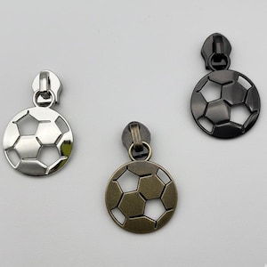 May include: Three metal zipper pulls in the shape of soccer balls. One is silver, one is bronze, and one is dark gray. The soccer ball design features a series of pentagons and hexagons.