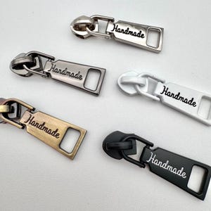 May include: A selection of metal zipper pulls in silver, gunmetal, gold, white, and black. Each pull is inscribed with the word "Handmade" in a cursive script. These pulls are for sewing and craft projects.