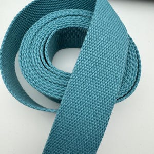 May include: A roll of teal-colored woven webbing. The webbing is tightly woven with a textured surface and is rolled into a spiral shape. The color is a muted teal, and the material appears to be durable and strong.