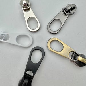 Puede incluir: Primer plano de cinco tiradores de cremallera metálicos en varios colores: plateado, blanco, negro y latón antiguo. Cada tirador tiene un agujero de forma ovalada para una fácil fijación. Estos tiradores son ideales para ropa, bolsos y otros proyectos de costura.