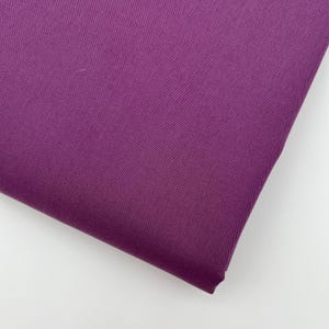 Op de afbeelding: Een close-up van een gevouwen stuk stof in een diepe paarse tint. De stof heeft een fijne, getextureerde weefsel en de plooien creëren schaduwen die de diepte en textuur van het materiaal benadrukken. De achtergrond is effen wit.