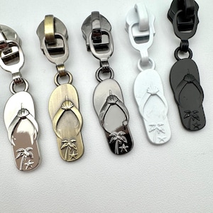 May include: Five metal zipper pulls in silver, bronze, gunmetal, white, and black. Each pull features a flip-flop charm with a palm tree and star design, perfect for adding a beachy touch to any project.