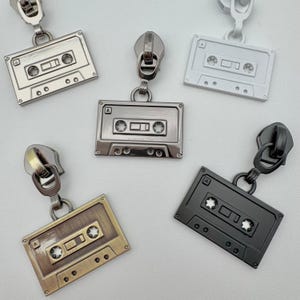 May include: A collection of cassette tape zipper pulls in various metallic finishes: silver, bronze, black, and white. Each pull features a detailed cassette tape design with a zipper pull attached at the top, perfect for adding a retro touch to bags or clothing.
