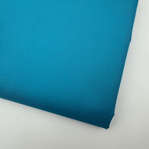 May include: A close-up shot of a folded piece of teal fabric. The fabric has a smooth texture and a slight sheen, with a visible diagonal weave pattern. The color is a vibrant, solid teal, with no other colors present.