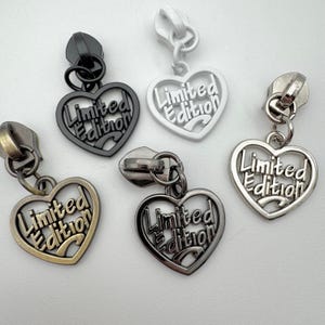 May include: A collection of heart-shaped zipper pulls in various metallic finishes: black, white, silver, bronze, and gunmetal. Each heart features the words "Limited Edition" in a stylized font, attached to a zipper.