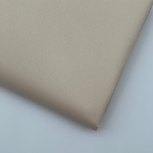 Op de afbeelding: Een gevouwen stuk beige stof. De stof heeft een subtiele textuur en een matte afwerking. De hoek van de stof is zichtbaar en toont de dikte en de manier waarop deze vouwt. De kleur is een neutraal, lichtbeige.