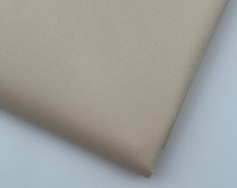 May include: A folded piece of beige fabric. The fabric has a subtle texture and a matte finish. The corner of the fabric is visible, showing the thickness and the way it folds. The color is a neutral, light tan.