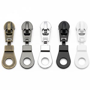 May include: Five metal zipper pulls in various colors: antique brass, gunmetal, silver, black, and white. Each pull has a rectangular top with a rounded pull tab and a circular hole at the bottom. These are ideal for clothing, bags, and craft projects.