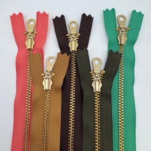 Puede incluir: Primer plano de cinco cremalleras abiertas en varios colores: coral, marrón, verde oliva y turquesa. Cada cremallera tiene un tirador y dientes de metal dorado. Las cremalleras están dispuestas verticalmente sobre un fondo blanco.