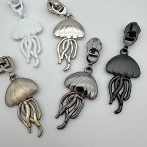 Può includere: Una collezione di tiretti per cerniera in metallo a forma di medusa. I tiretti sono in vari colori, tra cui bianco, argento e nero. Ogni tiretto presenta un design dettagliato di medusa con un cappuccio a cupola e tentacoli fluenti.