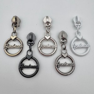 May include: Five metal zipper pulls in various finishes: antique brass, black, silver, and white. Each pull features a heart-shaped charm and a circular pendant with the word "Handmade" in script.