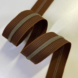May include: A spool of brown ribbon with a silver-colored chain trim. The ribbon has a ribbed texture with thin, dark brown stripes. A thin, white line runs down the center of the ribbon. The ribbon is curled and folded.