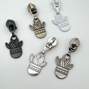 May include: A collection of decorative zipper pulls in various metallic finishes. Each pull features a stylized succulent plant design in a pot. Colors include black, silver, antique brass, and white. The pulls are designed for use in sewing and crafting projects.