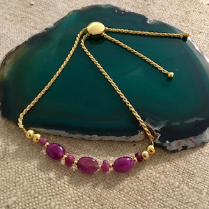 May include: A gold chain bracelet with a sliding clasp and purple oval-shaped beads. The bracelet is on a green agate surface.