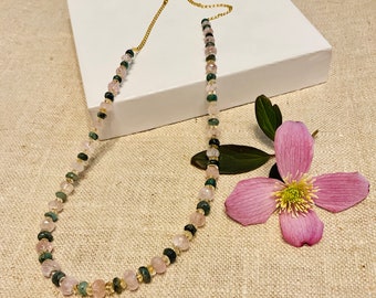 Könnte beinhalten: Eine zarte goldene Kettenkette mit abwechselnden rosa und grünen Edelsteinperlen. Die Kette liegt auf einer weißen Oberfläche mit einer rosa Blume.