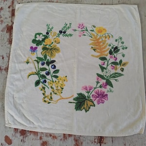 May include: A square off-white fabric napkin featuring a central wreath design composed of colorful botanical illustrations including yellow flowers, blue berries, green leaves, and pink blossoms, with the small initials G.L. printed in green.