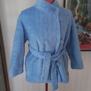 May include: A light blue, plush robe with a wrap-around design and a matching belt. The robe has long sleeves and a high collar. It is displayed on a mannequin against a white wall.