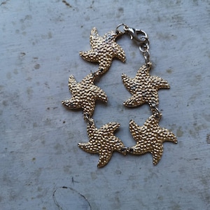 May include: Gold-toned bracelet with a chain and five star-shaped charms. The charms are textured and have a slightly raised design.
