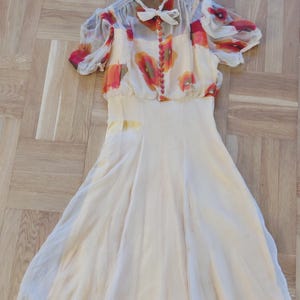 Könnte beinhalten: Ein cremefarbenes Vintage-Kleid mit floral bedrucktem Oberteil und transparenten Ärmeln. Das Oberteil hat eine zarte Schleife und kleine Knöpfe. Der fließende Rock schafft eine elegante Silhouette. Das Kleid hängt an einem Holzbügel.