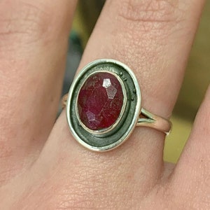 May include: A silver ring with a red gemstone set in a bezel setting. The ring is on a finger.