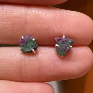May include: A pair of small stud earrings featuring raw ruby gemstones with a silver setting. The ruby stones display a mix of red and green hues. The earrings are held up by a finger.