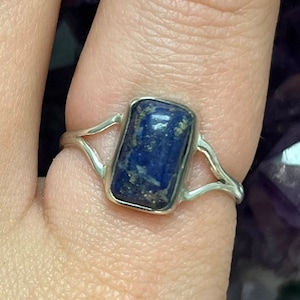 May include: A silver ring featuring a rectangular, deep blue gemstone with flecks of gold. The ring's band has a split design on either side of the stone, creating an open, airy look. The gemstone is set in a silver bezel.